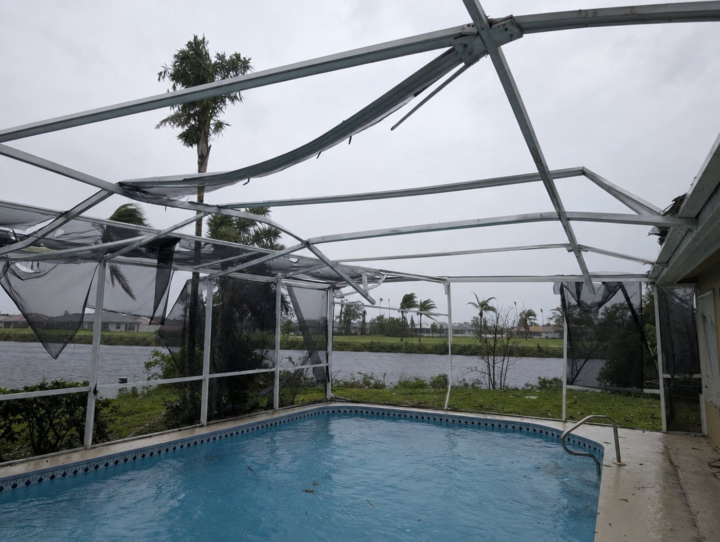 Storm damaged aluminum pool cage with a bent roof beam and missing screen panels in Florida