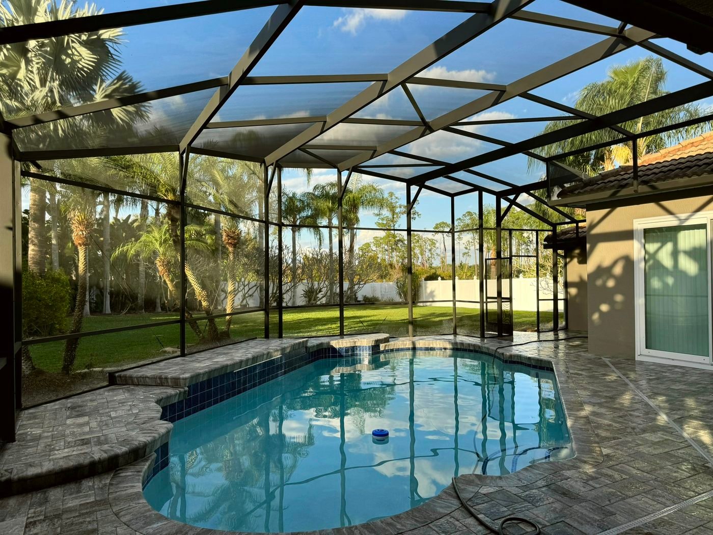 Dark bronze aluminum pool cage over a pool with a paver deck and palm trees beyond the screen