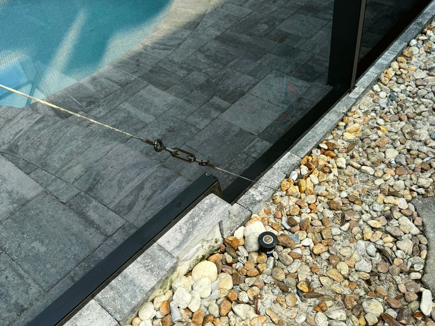 Dark bronze pool cage bottom rail along the edge of a paver deck, with a diagonal brace cable and turnbuckle beside a river rock border