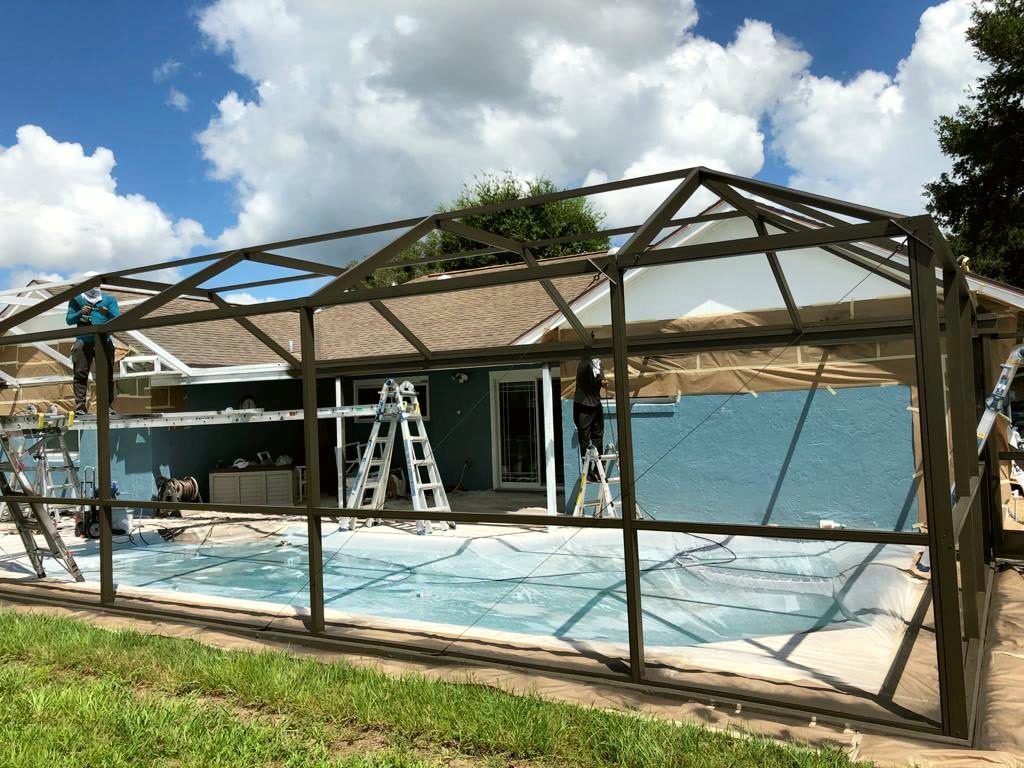 Crew on ladders working on a bare aluminum pool cage frame with the screens removed, the pool covered and the house wall masked