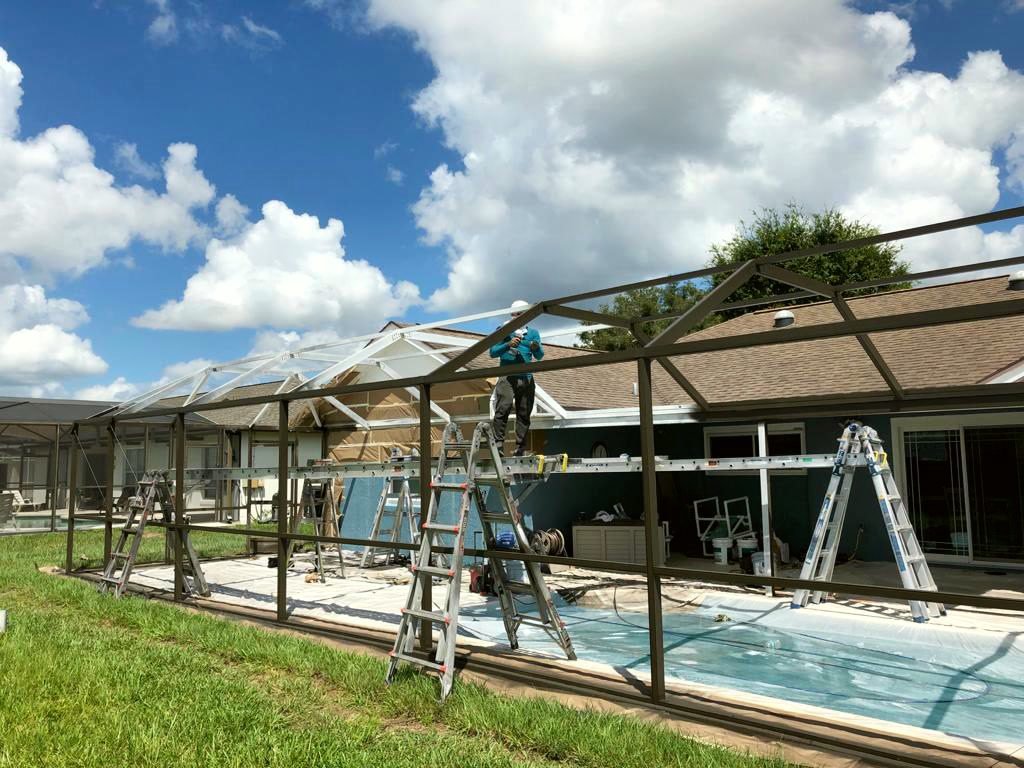 Crew member on a ladder working at the roof members of a pool cage frame, with the pool below covered in plastic