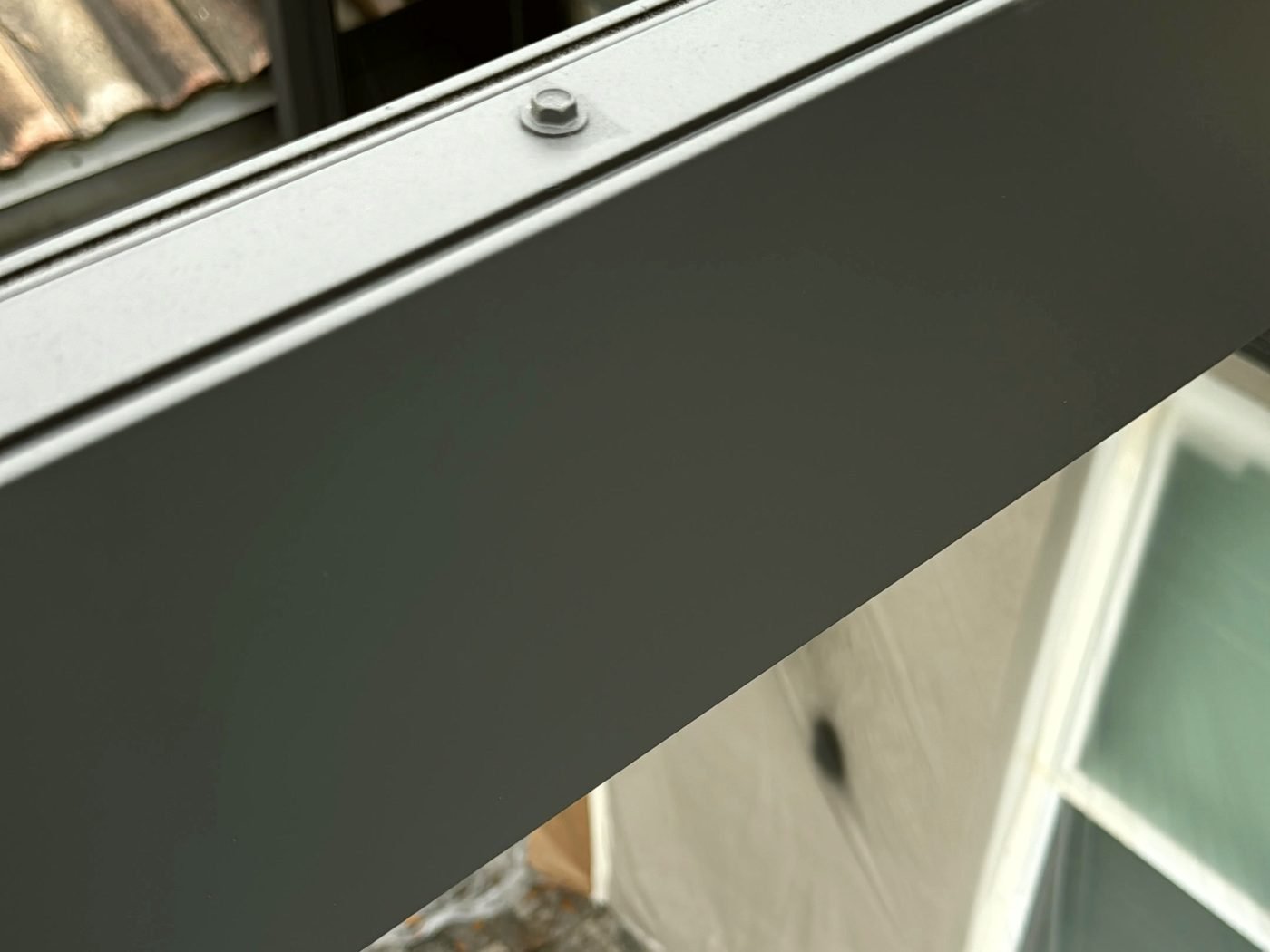 Close view of a painted dark pool cage frame member and a screw head, with a tile roof behind and the house wall below