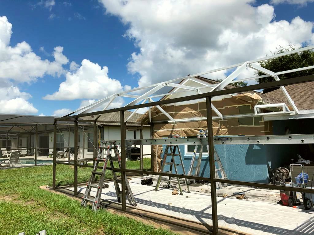Pool cage partway through a repaint, with bronze uprights and rails, white roof members, ladders inside and the house wall and deck covered